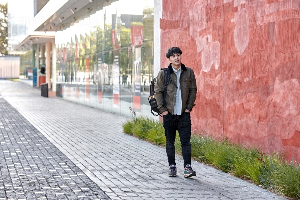 Young Asian university student walking on-campus next to red wall - Australian Stock Image
