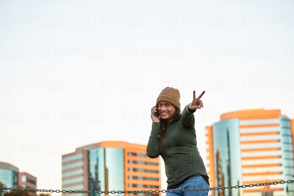 Young Asian woman doing peace sign with mobile phone in city - Australian Stock Image