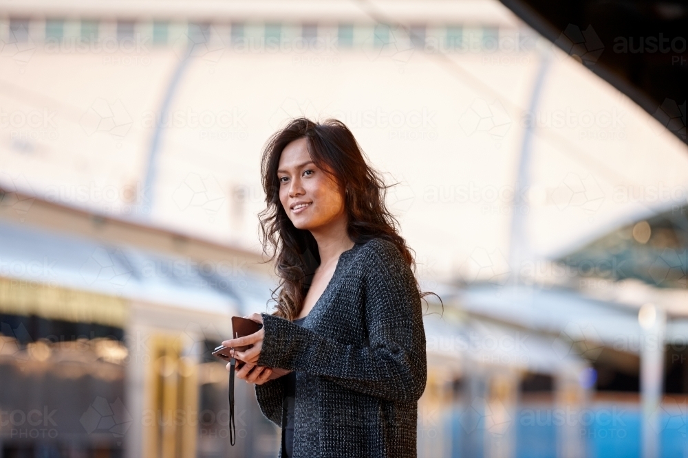 Young Asian woman waiting at train station with mobile phone - Australian Stock Image