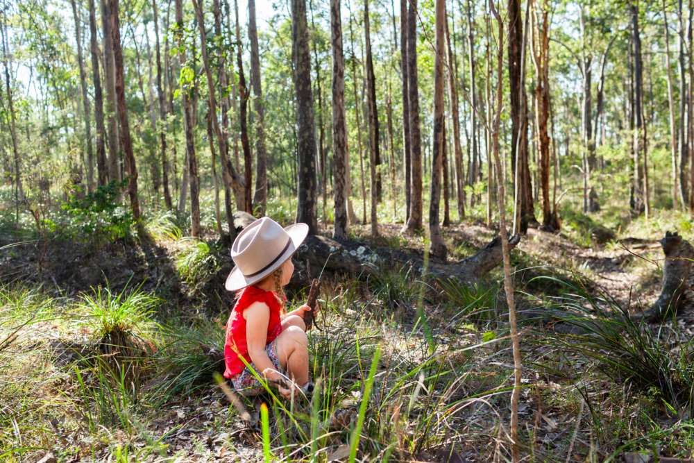 Young aussie kid wearing hat and red shirt sitting in australian bushland by fallen log - Australian Stock Image