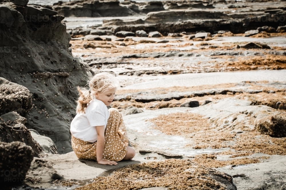 Young autistic girl sitting looking at rockpools at the beach - Australian Stock Image