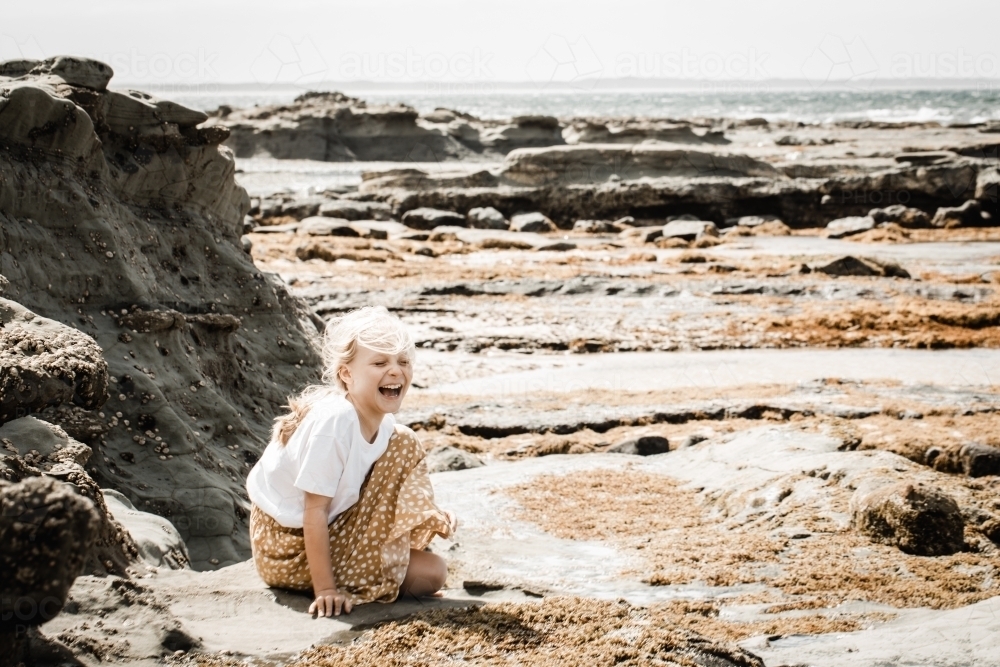 Young autistic girl sitting looking at rockpools at the beach laughing - Australian Stock Image