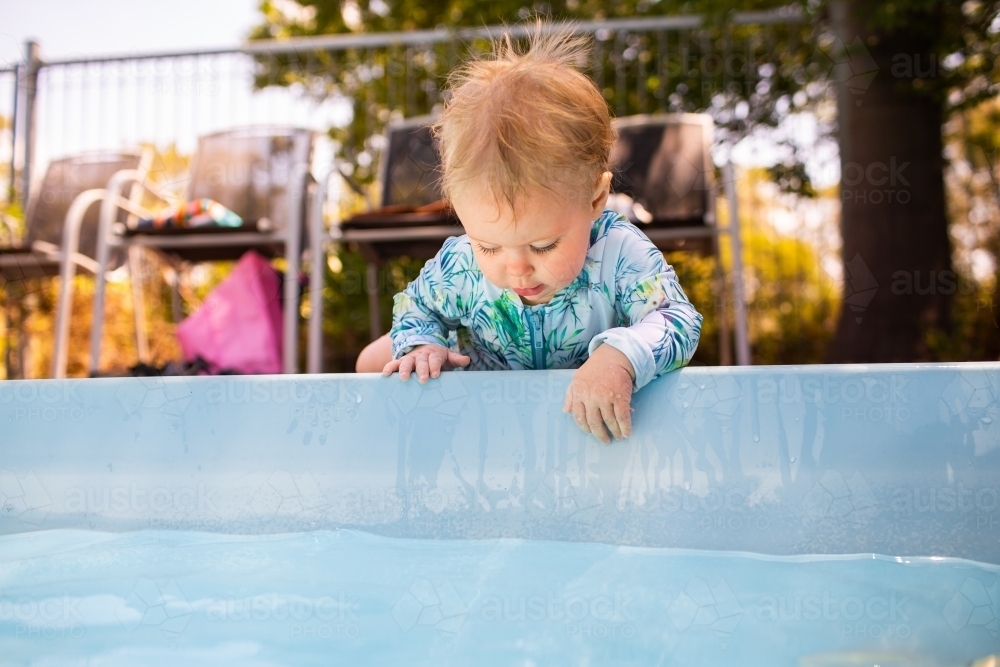 Young baby leaning over edge of inground swimming pool splashing in water – dangerous situation - Australian Stock Image