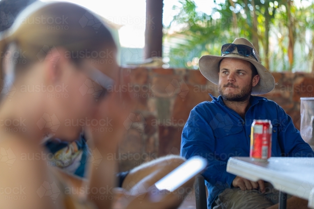 young bloke with cowboy hat, beer, sunnies with blurry foreground of girl on phone - Australian Stock Image