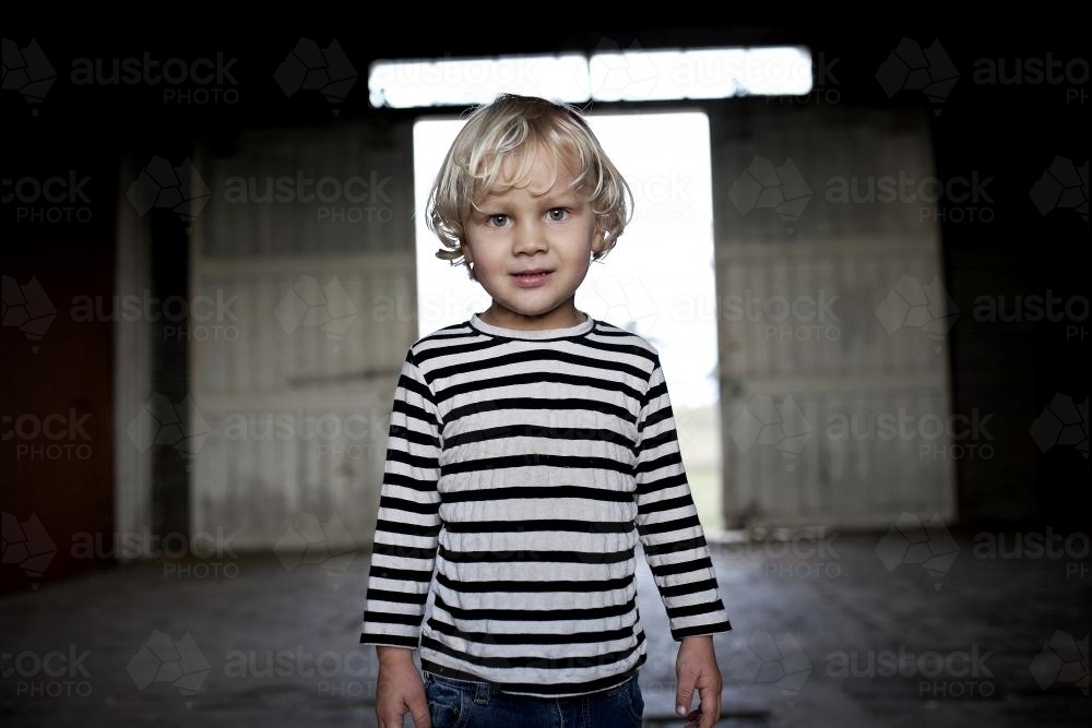 Young blond boy looking at camera with background of dimly lit shed - Australian Stock Image