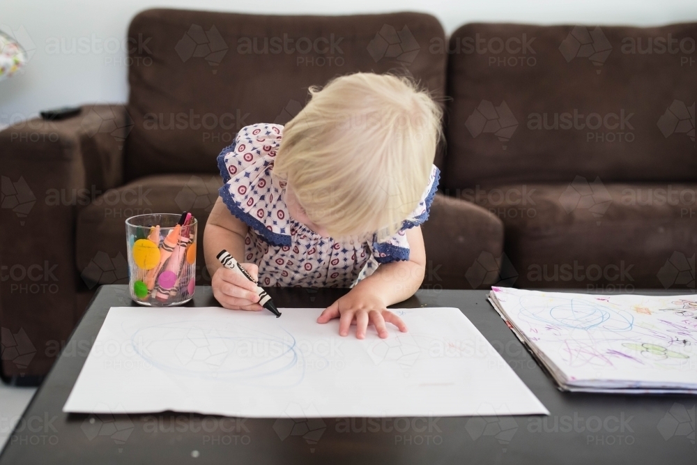 Young blonde girl bent over drawing on paper with crayons - Australian Stock Image