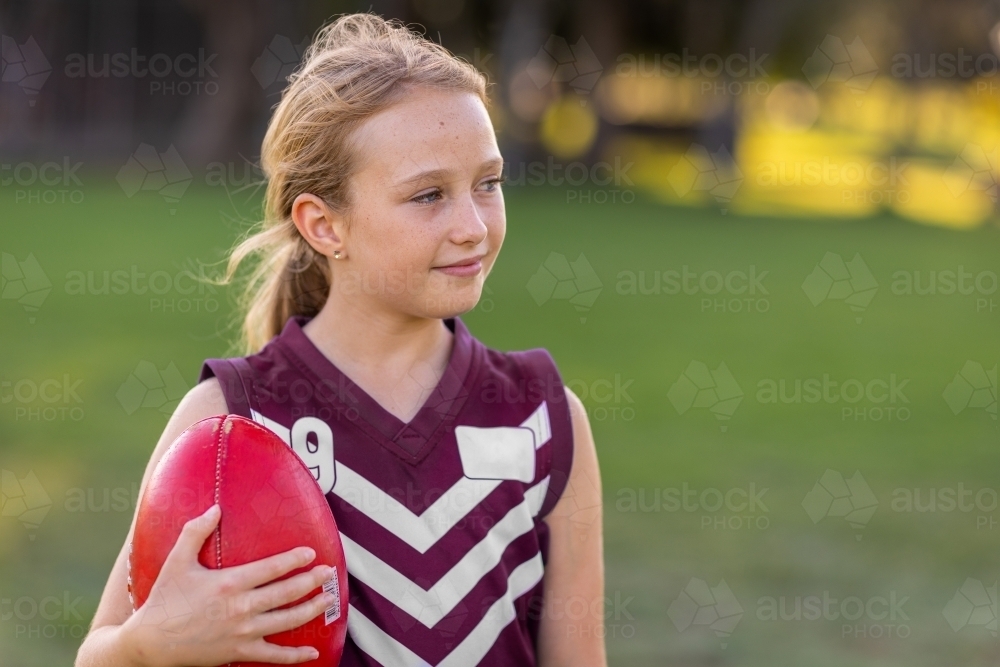young blonde girl holding red leather football and wearing a football jersey - Australian Stock Image