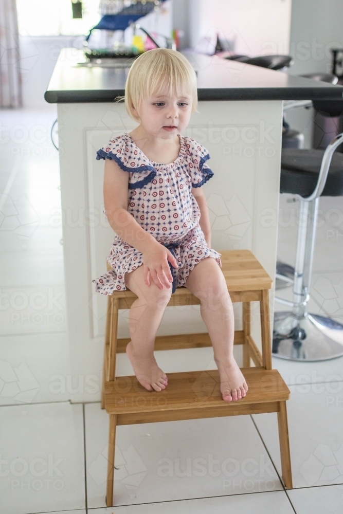 Young blonde girl sitting on step chair in kitchen at home - Australian Stock Image