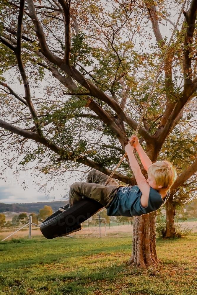 Young blonde hair boy playing on tire swing in country backyard - Australian Stock Image