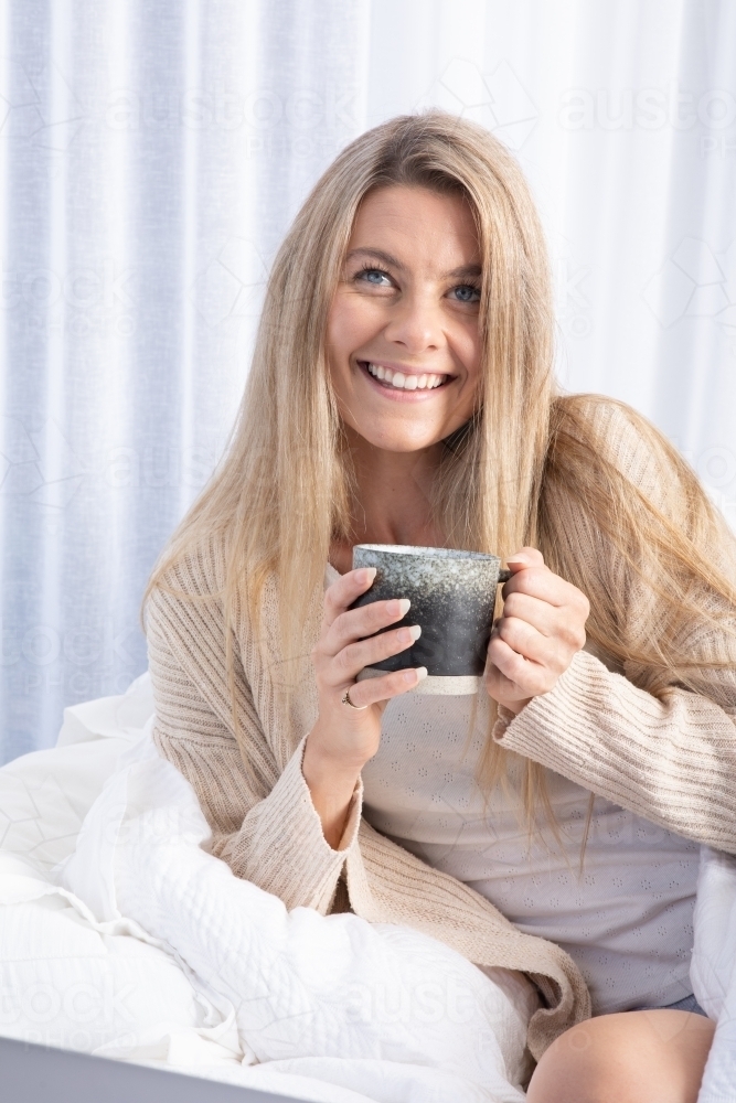 Young blonde lady smiling and drinking hot drink in white bed - Australian Stock Image