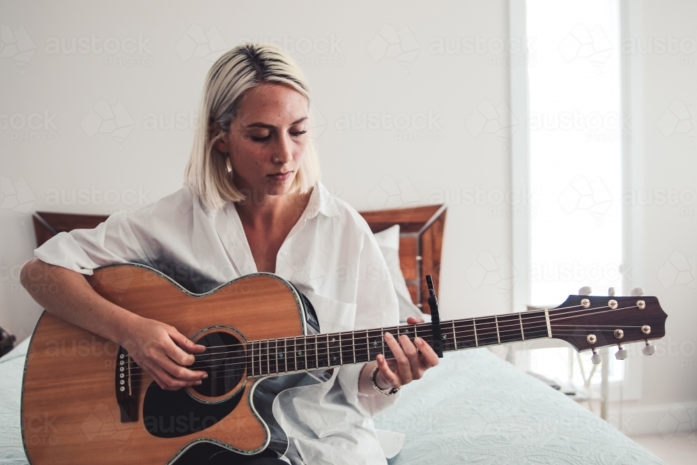 Young blonde woman in white sitting on bed playing guitar - Australian Stock Image
