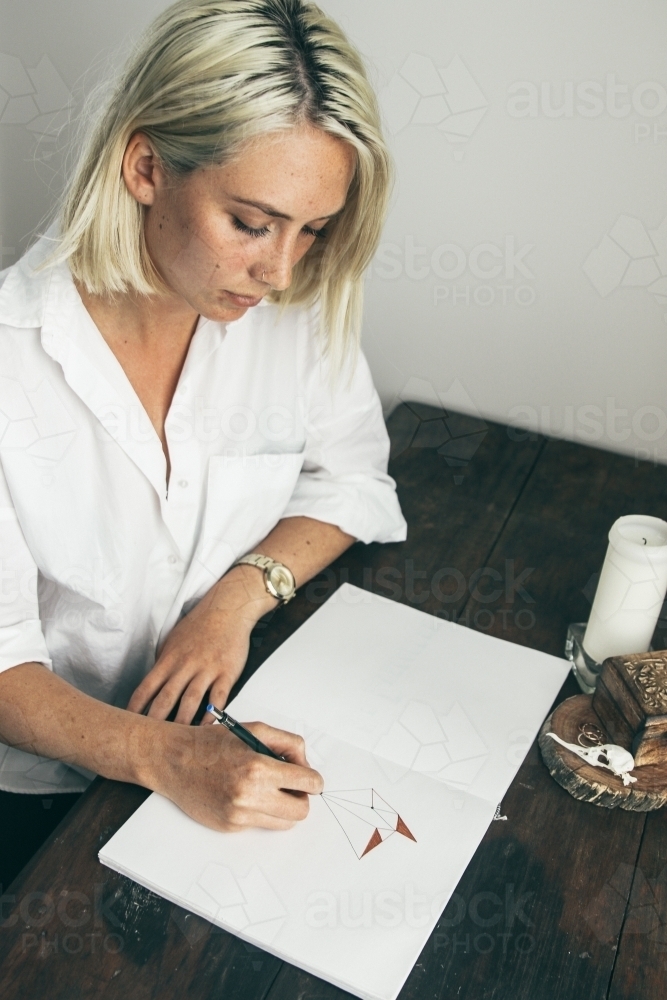 Young blonde woman sitting at desk drawing in a sketch book - Australian Stock Image