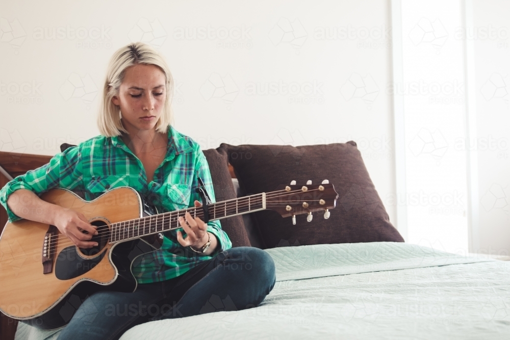 Young blonde woman sitting on bed playing guitar - Australian Stock Image