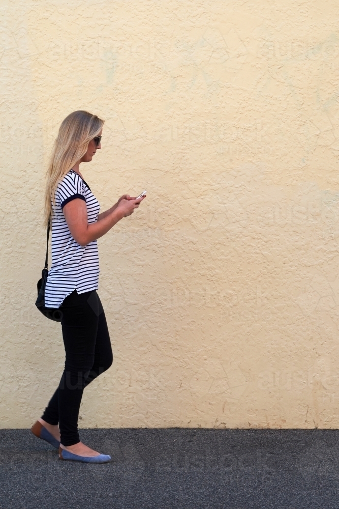 Young blonde woman walking and texting on mobile phone - Australian Stock Image