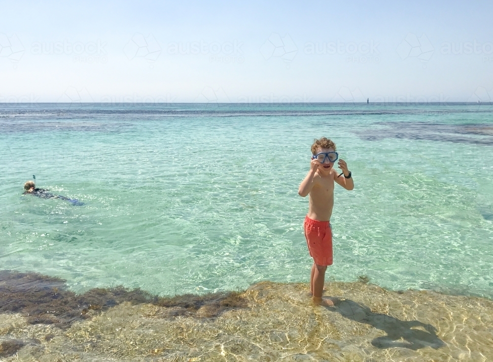 young boy adjusting mask on edge of clear lagoon standing on reef in orange board shorts - Australian Stock Image