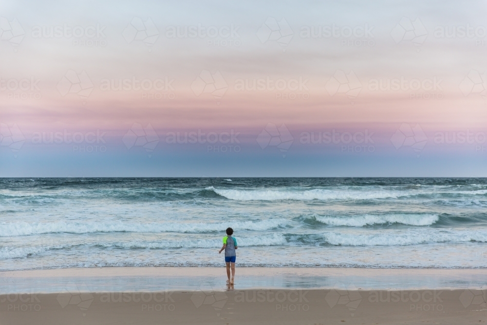 Young boy alone on beach at sunset looking at waves - Australian Stock Image