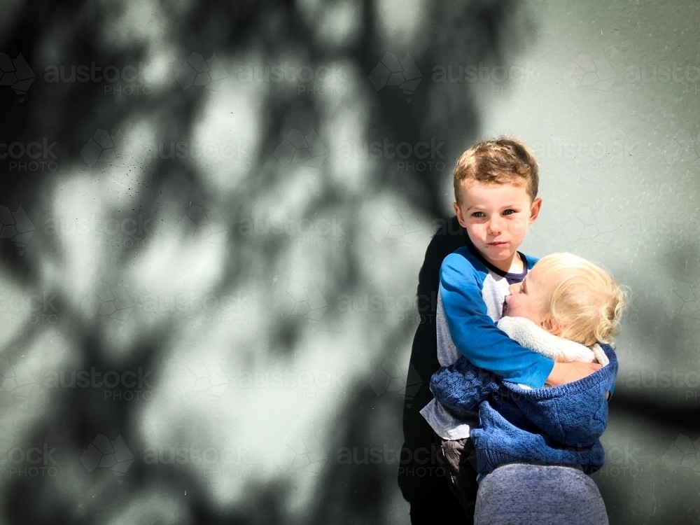 Young boy and toddler embracing against wall with tree shadowing them - Australian Stock Image
