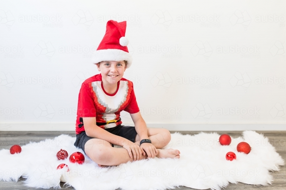 Young boy at Christmas wearing santa hat sitting on rug with baubles - Australian Stock Image