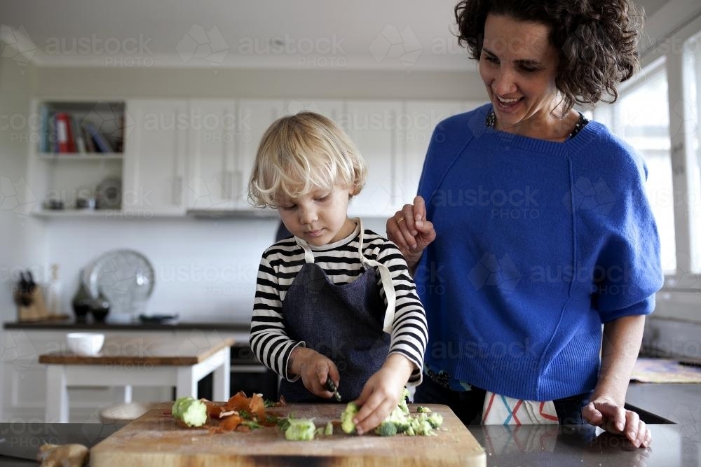 Young boy being supervised by his mum while chopping veggies - Australian Stock Image