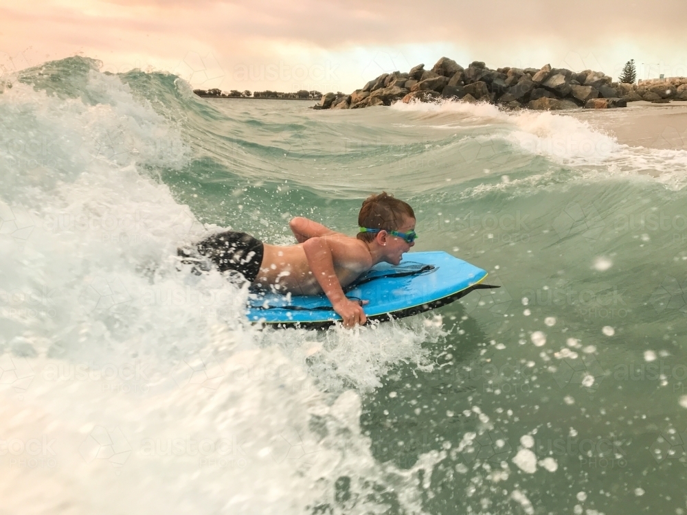 Young boy catching a wave on his bodyboard with rock groyne in background - Australian Stock Image