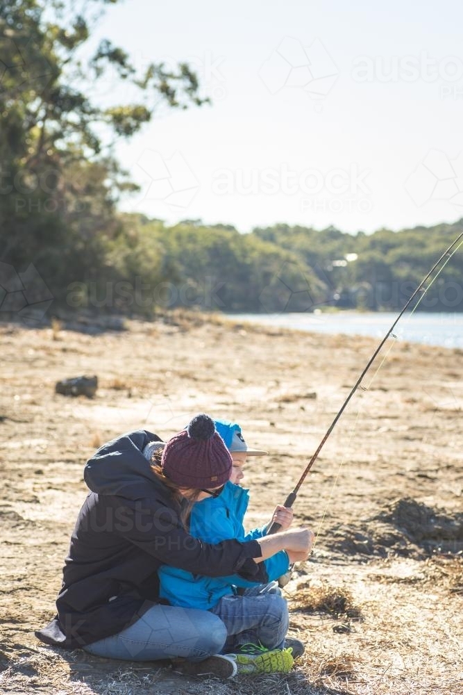 Young boy cuddling with mother while fishing on the banks of a river - Australian Stock Image