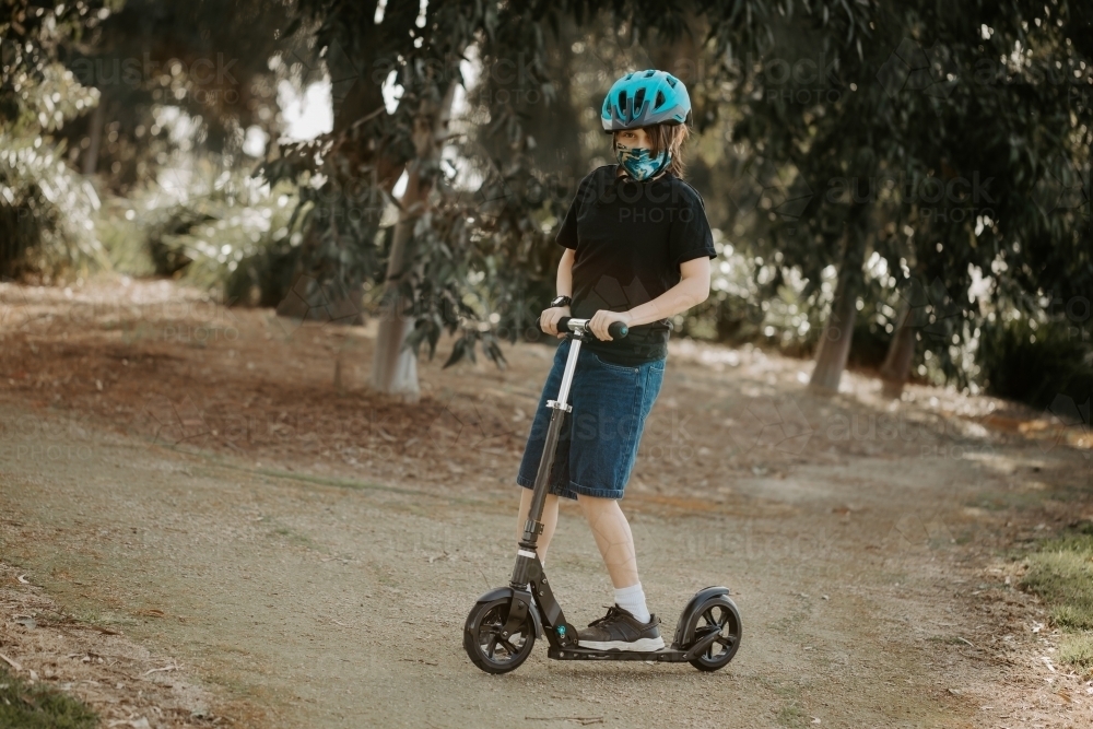 Young boy during the corona virus COVID-19 pandemic wearing a fabric mask and riding scooter - Australian Stock Image