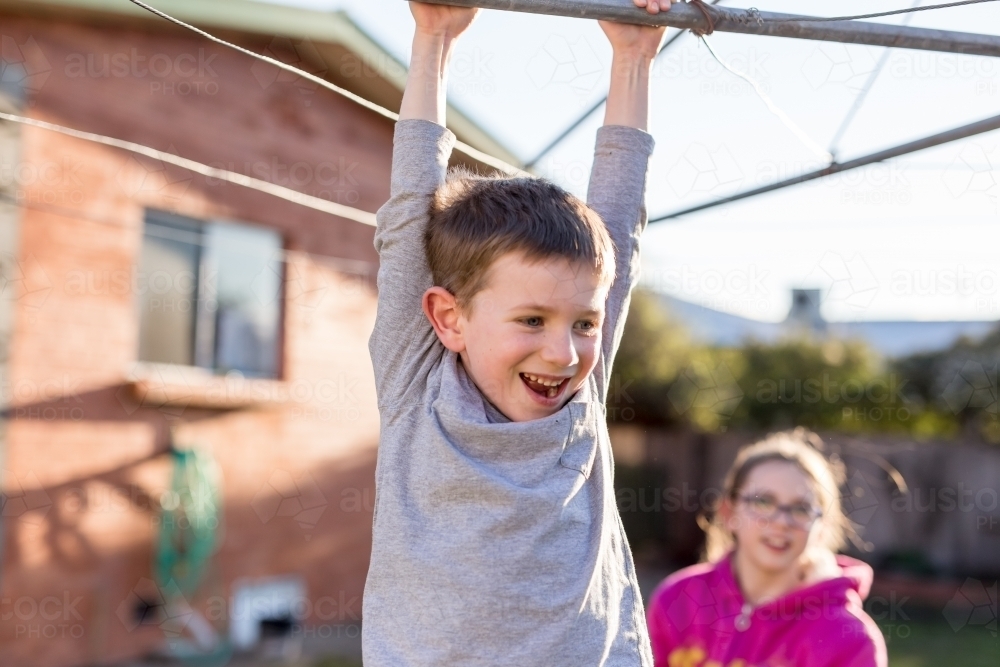 Young boy hanging from washing line being chased by sister - Australian Stock Image