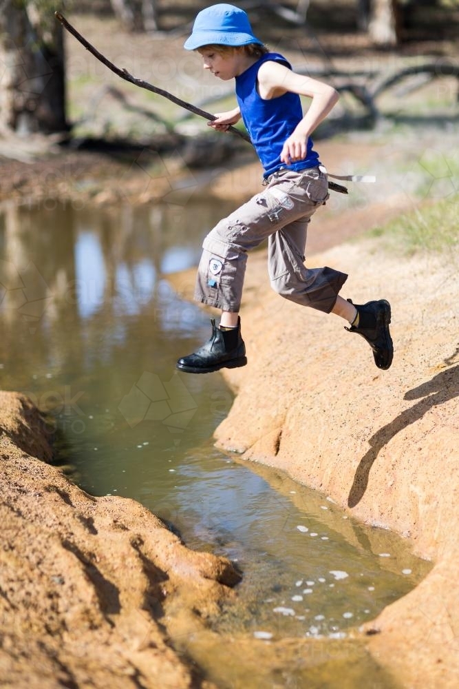 young boy holding a stick while jumping across a creek - Australian Stock Image