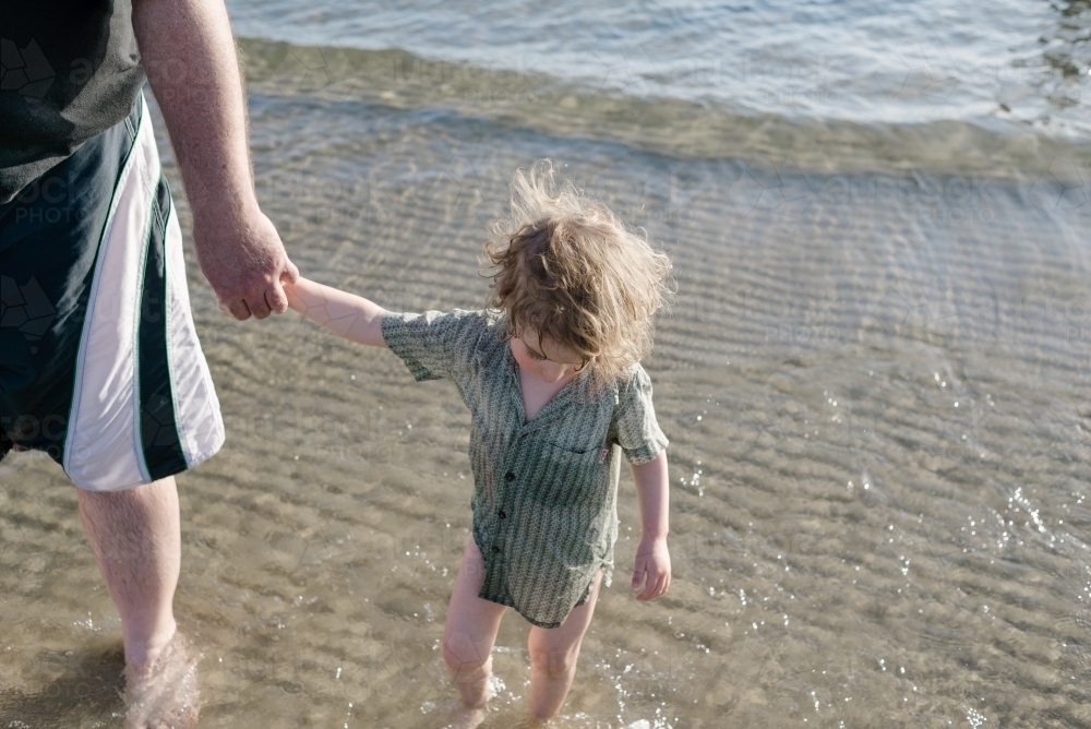 Young boy holding his father's hand while walking in the ocean - Australian Stock Image
