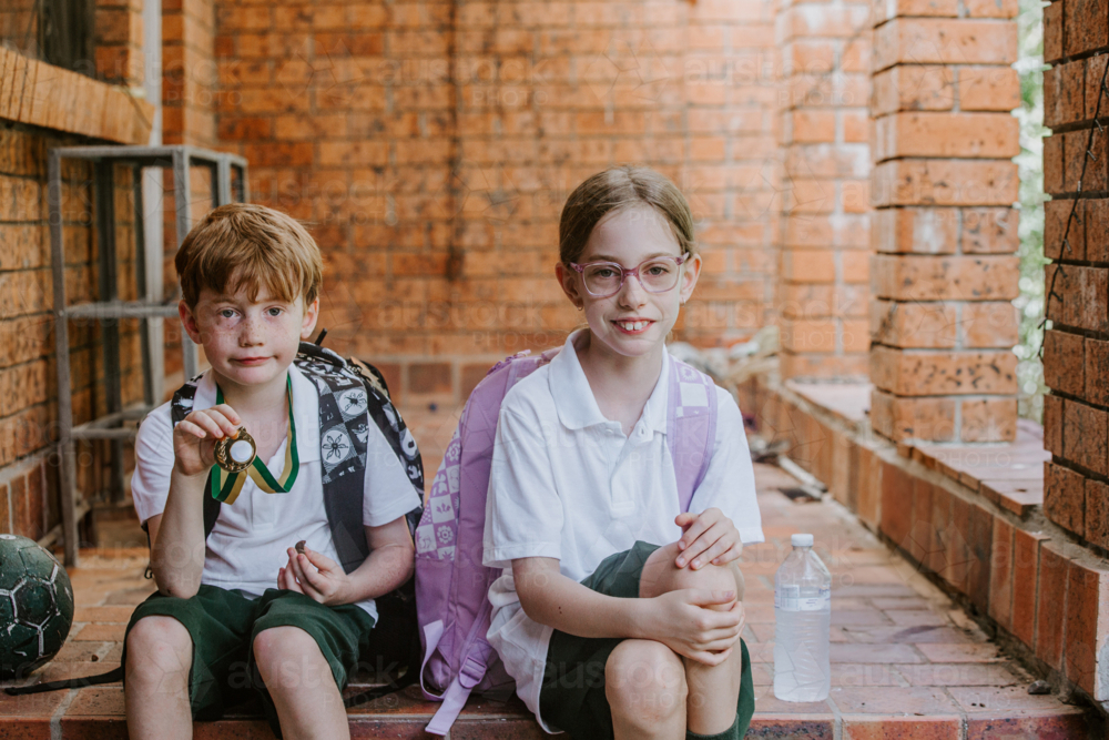 Young boy holding his medal sitting on the brick floor of a house. with sister - Australian Stock Image