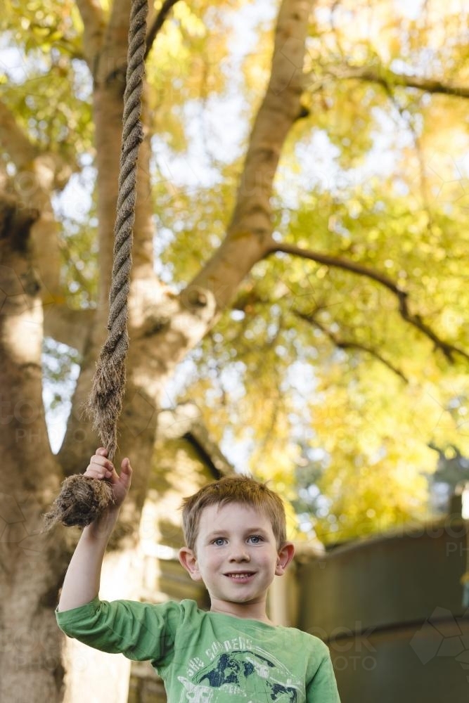 Young boy holding onto a swinging rope under tree - Australian Stock Image