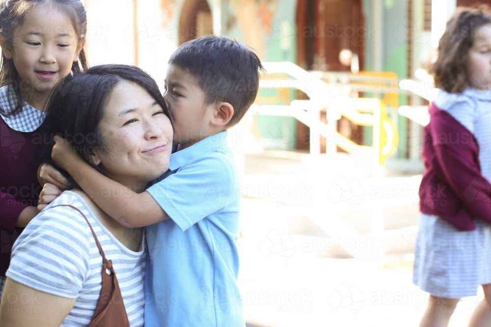 Young boy in school uniform hugging his mum while his sister looks on - Australian Stock Image