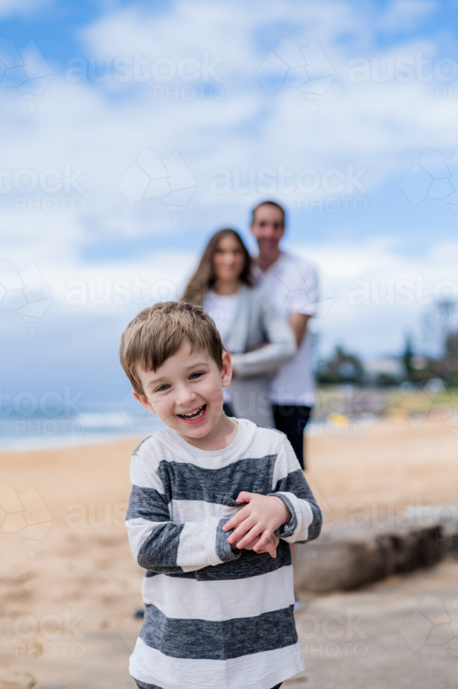 Young boy joyfully smiles while parents stand behind him at the beach - Australian Stock Image