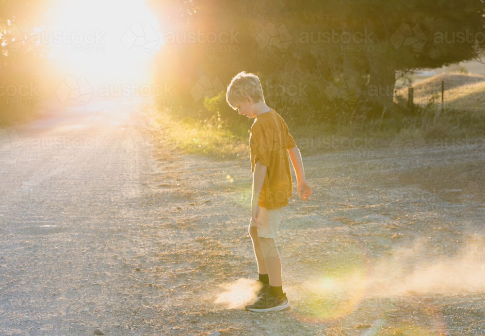 young boy kicking dirt on side of country road at sunset - Australian Stock Image