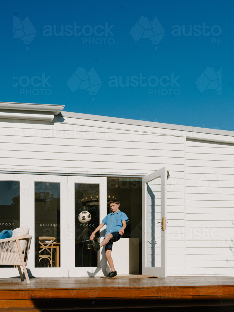 Young boy kicking soccer ball on garden deck of white weatherboard home - Australian Stock Image