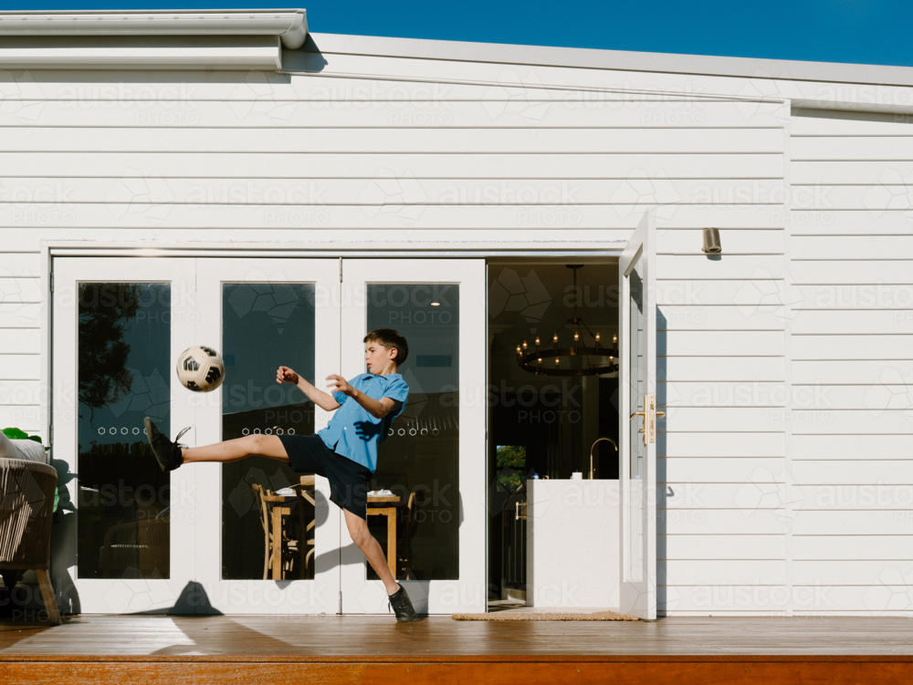 Young boy kicking soccer ball on their garden deck. - Australian Stock Image