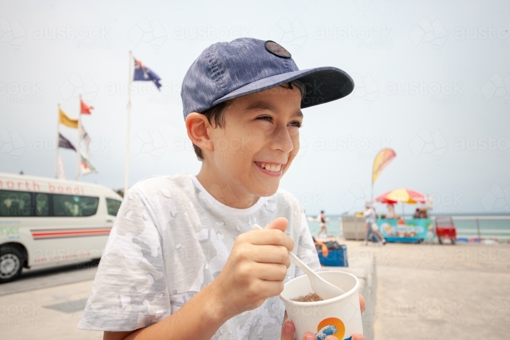 Young boy laughing whilst eating snow cone at beach - Australian Stock Image