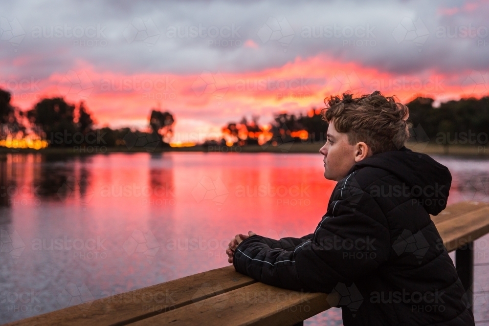 Young boy leaning on wooden railing looking out over river water at sunset - Australian Stock Image