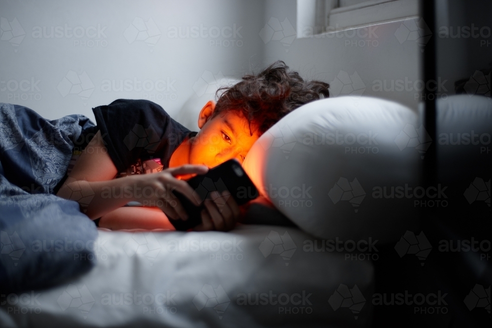 Young boy looking at device in bed - Australian Stock Image