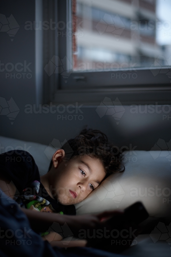 Young boy looking at device in bed - Australian Stock Image