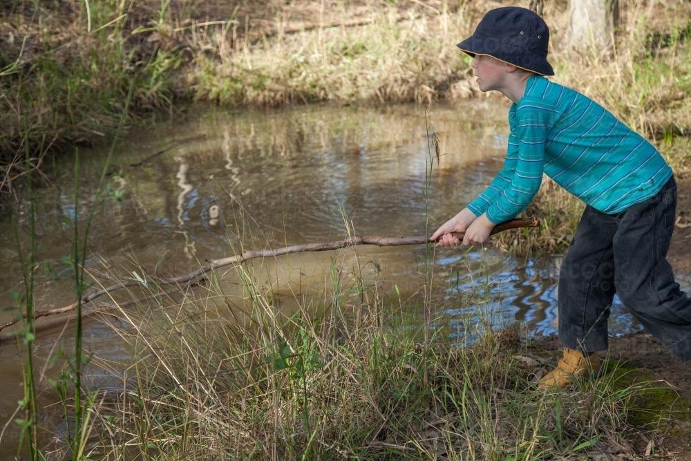 Young boy playing with a stick at the creek - Australian Stock Image