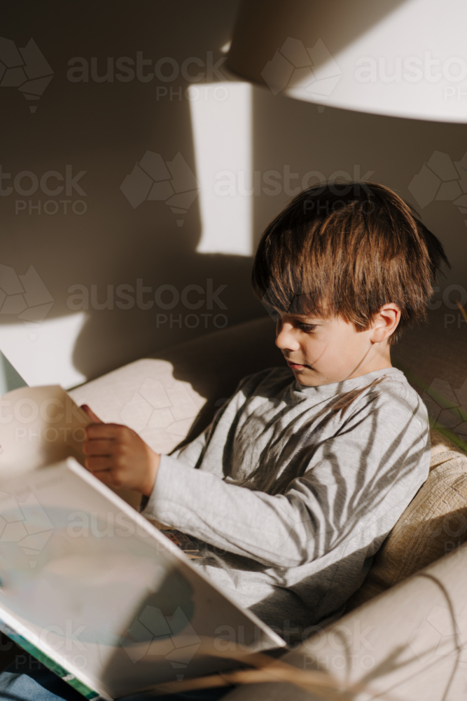 Young boy reading a book on the couch in a sunlit room. - Australian Stock Image