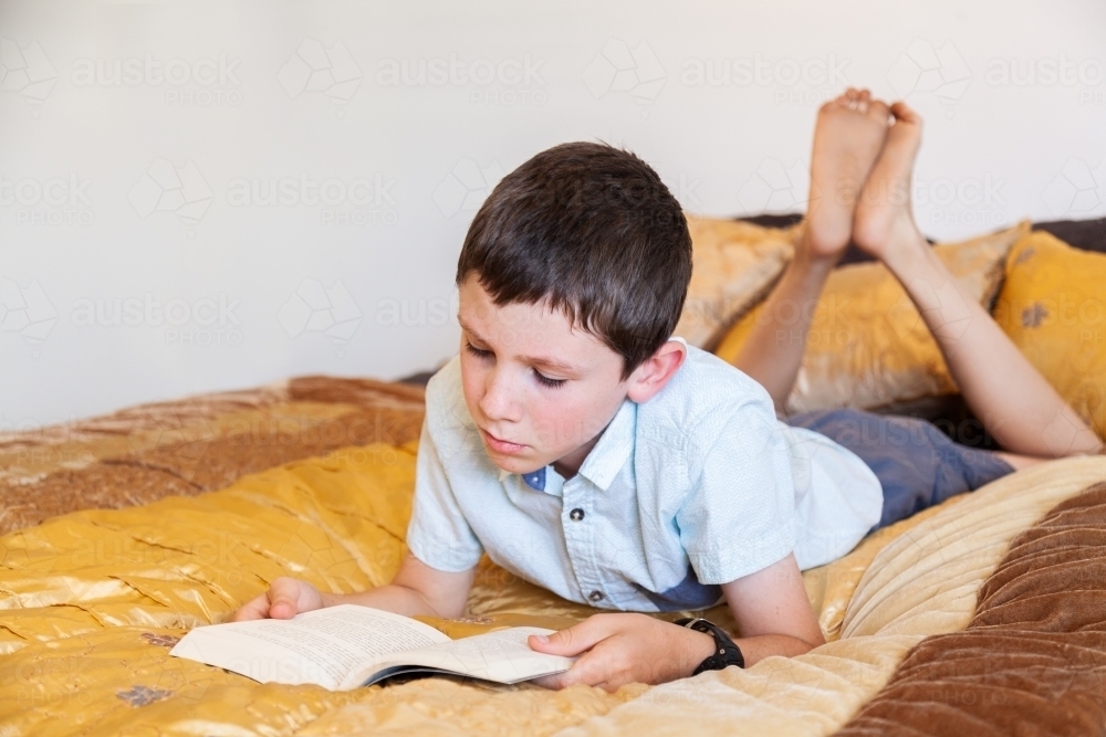 Young boy reading fiction book on bed in bedroom - Australian Stock Image