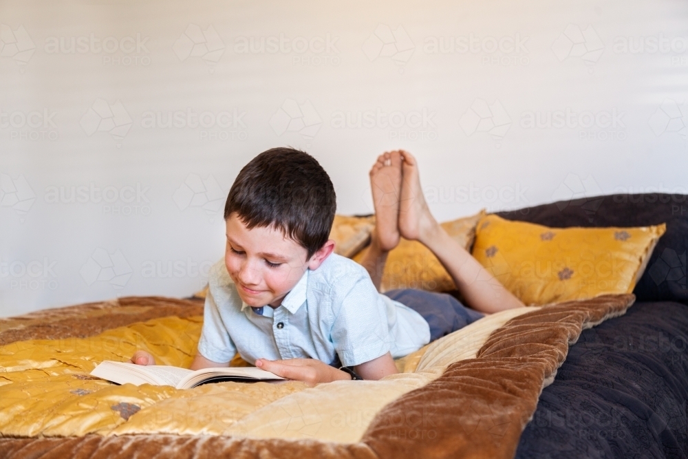 Young boy reading fiction book on bed in bedroom - Australian Stock Image