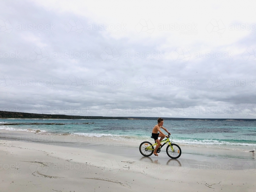 young boy riding his bike on empty beach on overcast summers day - Australian Stock Image