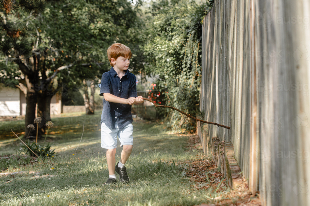Young boy running a piece of stick along a wooden fence - Australian Stock Image
