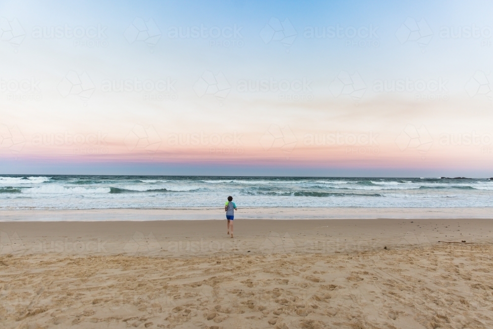 Young boy running from sand towards waves on beach at sunset - Australian Stock Image
