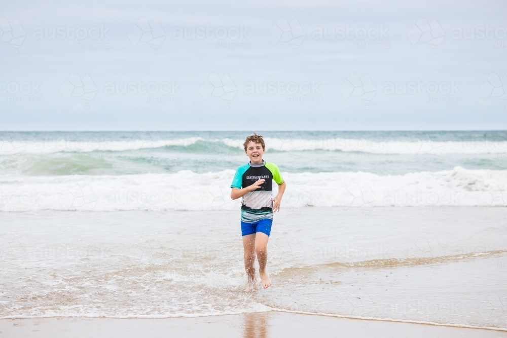 Young boy running in waves on beach happy smiling - Australian Stock Image