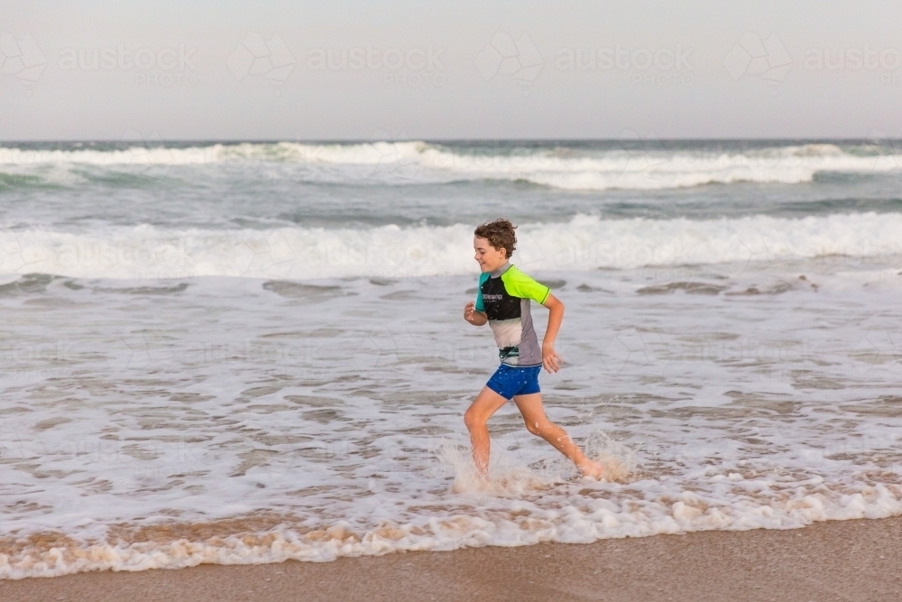 Young boy running through water on beach in afternoon - Australian Stock Image