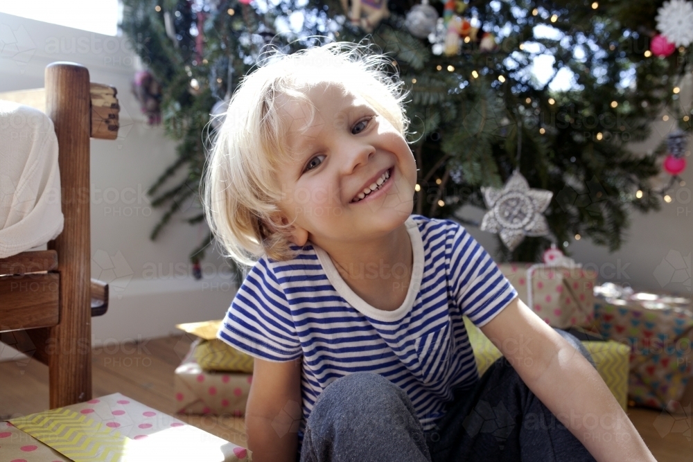 Young boy sitting in front of Christmas tree looking and smiling at camera - Australian Stock Image